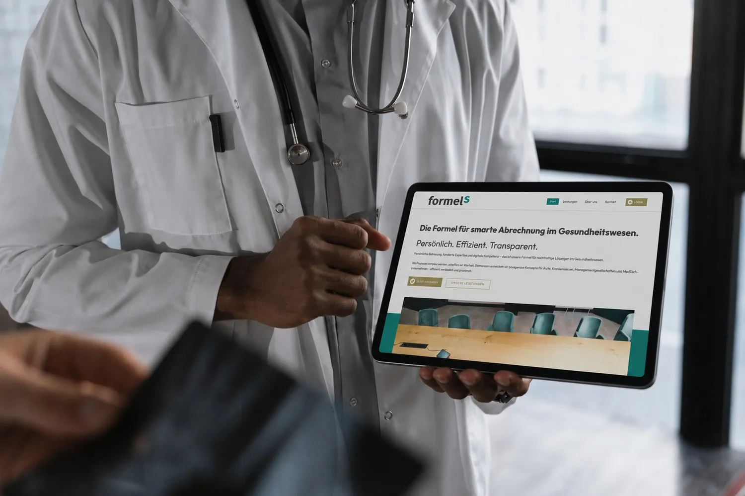 formelS GmbH – Berufsbildungszentrum Halle – Ein Arzt in weißem Kittel mit einem Stethoskop hält ein Tablet in der Hand, während er sich in einem hellen Krankenhauszimmer mit jemandem unterhält.