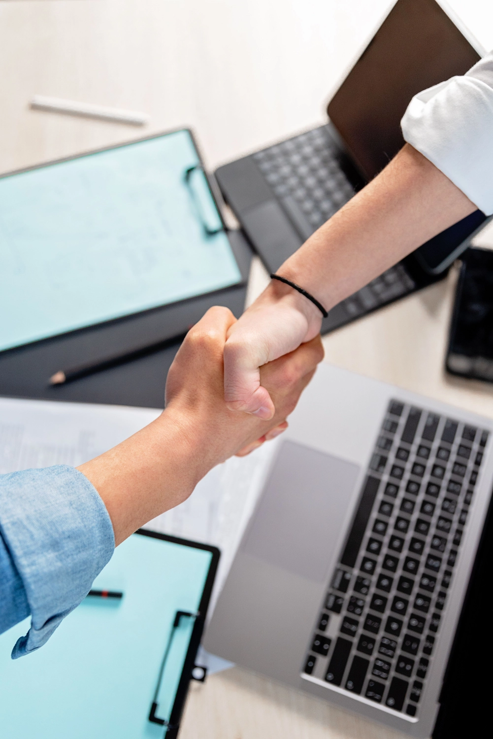formelS GmbH – Berufsbildungszentrum Halle – Zwei Personen geben sich über einem Schreibtisch mit Laptops, Schreibunterlagen und Unterlagen die Hand, was auf eine geschäftliche Vereinbarung hindeutet.